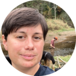 A person with short dark hair smiles at the camera outdoors near a stream, with a dog and another person in the background surrounded by greenery.