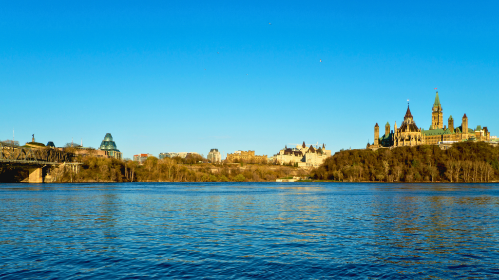 Scenic view of a calm river with the Canadian Parliament buildings, including the iconic Peace Tower, perched on a hill under a clear blue sky, with surrounding historic architecture and greenery.