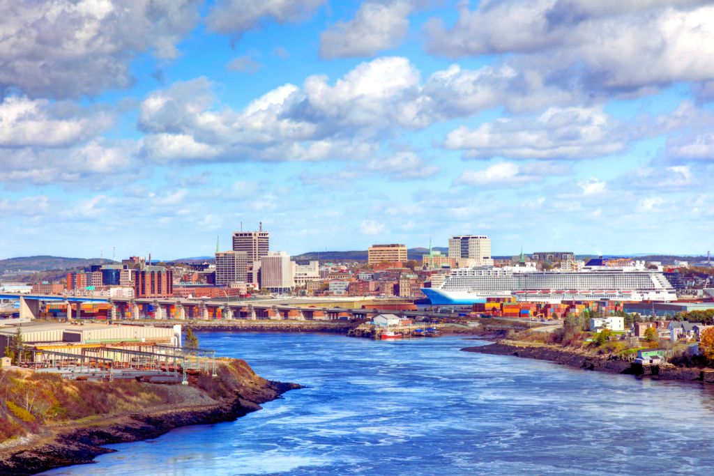 Vibrant cityscape view of a waterfront city with modern buildings, a large cruise ship docked at the port, and a flowing river in the foreground under a bright, partly cloudy sky.