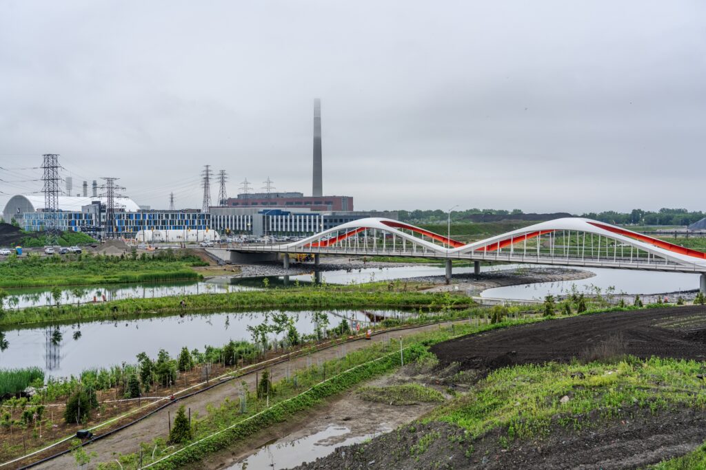 An urban landscape featuring a modern, wavy pedestrian bridge with red and white accents spanning over a series of wetlands, surrounded by newly planted greenery, with industrial buildings and power lines in the background under a cloudy sky.